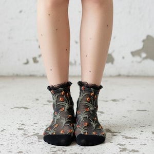May include: Sheer black ankle socks with a floral pattern in shades of orange and green. The socks have a black ruffled trim at the top and solid black toes.