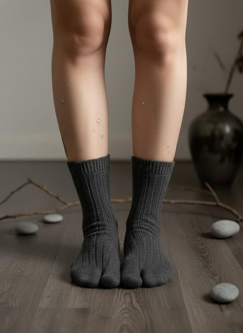 Puede incluir: Calcetines tabi de color gris oscuro con una textura acanalada. Los calcetines tienen un dise&ntilde;o de puntera dividida y se usan sobre un suelo de madera. La imagen muestra las piernas y los pies inferiores, con un fondo neutro.