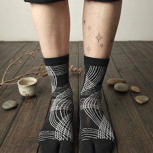May include: Black tabi socks featuring a white, wavy line pattern. The socks have a split toe and reach mid-calf. The image shows the socks on a person's legs, with a wooden floor in the background. The person has star tattoos on their leg.