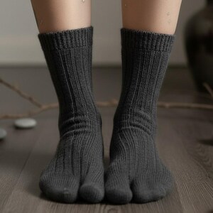 Puede incluir: Un par de calcetines tabi de color gris oscuro, de punto acanalado, con un dise&ntilde;o de puntera dividida. Los calcetines se llevan sobre un suelo de madera. La imagen muestra las piernas y los pies. Los calcetines son de color s&oacute;lido.