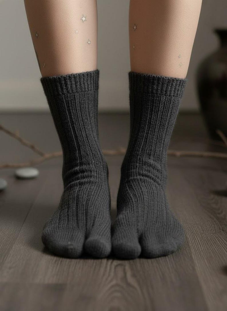 Puede incluir: Un par de calcetines tabi de color gris oscuro, de punto acanalado, con un dise&ntilde;o de puntera dividida. Los calcetines se llevan sobre un suelo de madera. La imagen muestra las piernas y los pies. Los calcetines son de color s&oacute;lido.