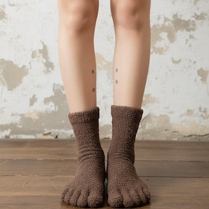 Puede incluir: Un par de calcetines de dedos marrones y difusos. Los calcetines se usan en los pies de una persona, con los dedos separados. El fondo es una pared de color claro y un suelo de madera.