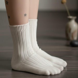 Puede incluir: Un par de calcetines acanalados de color crema. Los calcetines est&aacute;n hechos de un material suave y tienen un dise&ntilde;o cl&aacute;sico. Los calcetines se muestran en las piernas de una persona. El fondo est&aacute; borroso, con un jarr&oacute;n y un suelo de madera oscura.