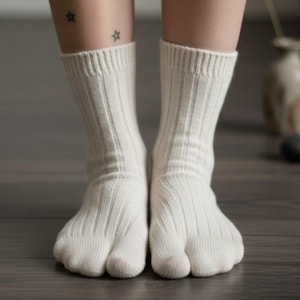 Puede incluir: Un par de calcetines blancos de punto acanalado con un dise&ntilde;o de puntera dividida. Los calcetines se usan en los pies de una persona, con las piernas y los pies inferiores visibles. El fondo es un suelo de madera.