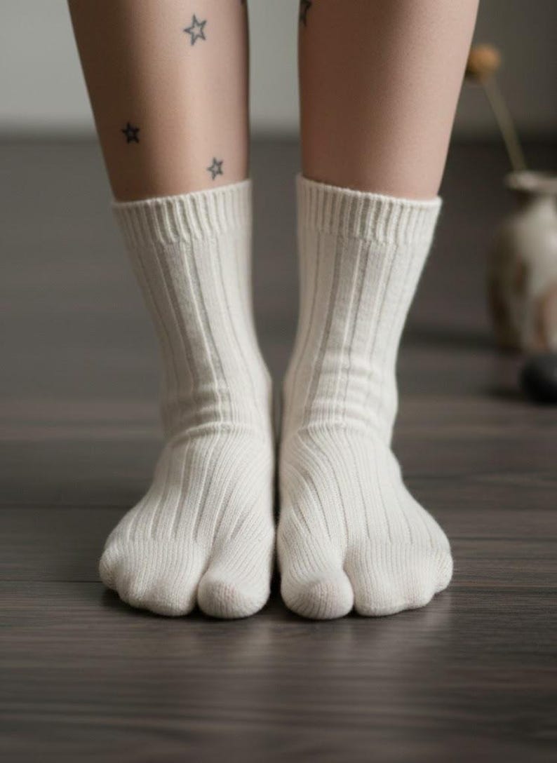 Puede incluir: Un par de calcetines blancos de punto acanalado con un dise&ntilde;o de puntera dividida. Los calcetines se usan en los pies de una persona, con las piernas y los pies inferiores visibles. El fondo es un suelo de madera.