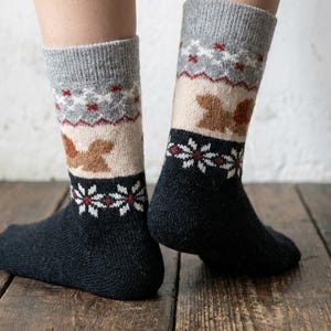 Puede incluir: Un par de calcetines estampados con un puño gris, una sección beige con figuras de ardillas marrones y un pie azul marino. Patrones florales blancos y rojos adornan el diseño. Los calcetines se muestran sobre una superficie de madera.