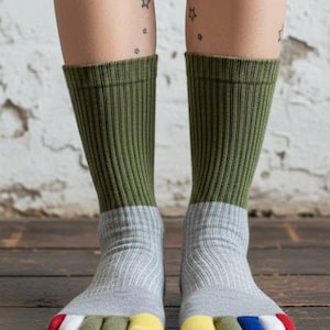 May include: A pair of toe socks with a color-blocked design. The socks feature olive green cuffs, gray midsections, and individual toe compartments in red, white, green, yellow, and blue. The socks are ribbed and worn on a wooden surface.