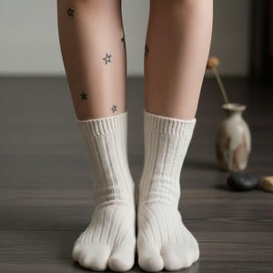 Puede incluir: Un par de calcetines de punto acanalado blanco roto con dise&ntilde;o de puntera dividida. Los calcetines se llevan en pies con tatuajes de estrellas en las piernas. Los calcetines est&aacute;n sobre un suelo de madera oscura.