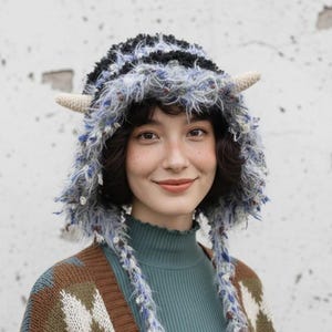 Puede incluir: Un sombrero artesanal caprichoso con un diseño peludo y texturizado en tonos de azul, gris y negro. El sombrero presenta dos pequeñas protuberancias en forma de cuerno de color crema. La persona también lleva un cuello alto turquesa y una rebeca estampada marrón y azul.