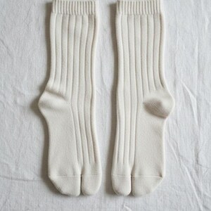 Puede incluir: Un par de calcetines color crema, acanalados, con dise&ntilde;o de puntera dividida. Los calcetines est&aacute;n hechos de un material suave y se muestran sobre una superficie blanca texturizada. Los calcetines tienen un estilo cl&aacute;sico y minimalista.