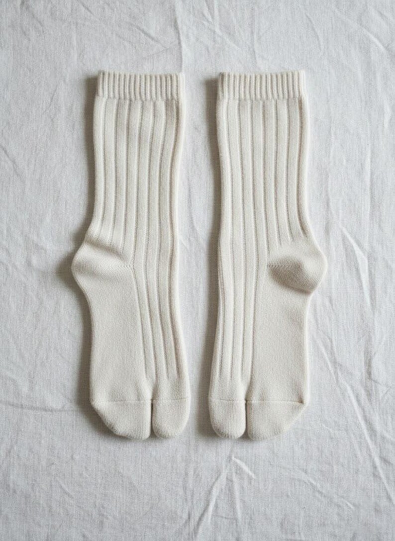 Puede incluir: Un par de calcetines color crema, acanalados, con dise&ntilde;o de puntera dividida. Los calcetines est&aacute;n hechos de un material suave y se muestran sobre una superficie blanca texturizada. Los calcetines tienen un estilo cl&aacute;sico y minimalista.