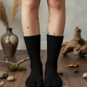 Puede incluir: Calcetines tabi negros, de punto acanalado, usados en un suelo de madera. Los calcetines tienen un dise&ntilde;o de puntera dividida. Las piernas tienen tatuajes de estrellas. El fondo incluye un jarr&oacute;n, plantas secas y piedras.