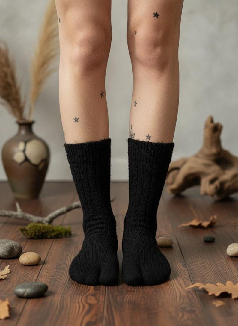 Puede incluir: Calcetines tabi negros, de punto acanalado, usados en un suelo de madera. Los calcetines tienen un dise&ntilde;o de puntera dividida. Las piernas tienen tatuajes de estrellas. El fondo incluye un jarr&oacute;n, plantas secas y piedras.