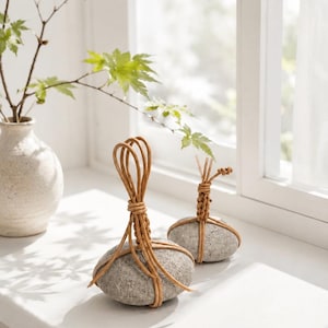 May include: Two smooth, grey, oval-shaped stones wrapped in thin, light brown, flexible wooden strips. The strips are intricately knotted and looped at the top of each stone, creating a decorative handle-like structure for these natural objects.