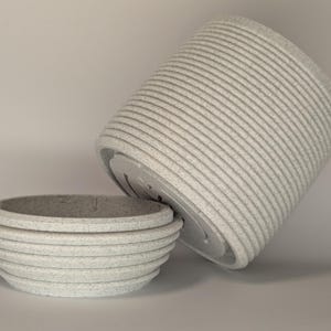 Op de afbeelding: Twee lichtgrijze, getextureerde keramische plantenbakken. Eén staat rechtop, de andere is gekanteld en onthult de drainagegaten. De plantenbakken hebben een geribbeld, horizontaal ontwerp. De kleinere plantenbak heeft een komvorm.