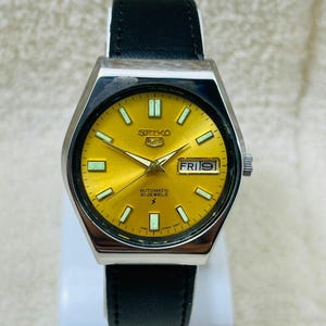 May include: A vintage Seiko automatic wristwatch with a gold-colored dial and a black leather strap. The watch face displays the day and date, with luminous markers and hands. The watch has a silver-colored, octagonal case.