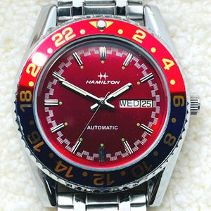 Puede incluir: Un reloj de pulsera automático Hamilton de acero inoxidable con esfera roja y bisel giratorio. El bisel presenta secciones rojas y azules con números blancos. La esfera muestra el día y la fecha, con la palabra "AUTOMATIC" impresa debajo.