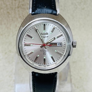 May include: Silver wristwatch with a black leather strap. The watch face is silver with black and green markers, a red second hand, and the day and date displayed. The watch has the text "hmt Rajat" and "AUTOMATIC 21 JEWELS".