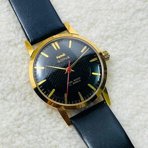 May include: A vintage wristwatch with a black patterned face, gold bezel, and a black leather strap. The watch face has gold hour markers and hands, with a red second hand. The watch is marked "Sona" and "Para Shock 17 Jewels".