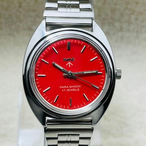 May include: A silver-toned wristwatch with a vibrant red face. The watch features a silver band and a round face with black hands and markers. The text "PARA SHOCK 17 JEWELS" is displayed on the watch face.
