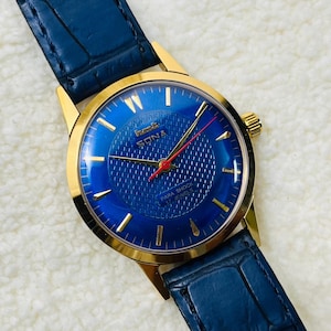 May include: A gold wristwatch with a vibrant blue face and a blue leather strap. The watch face displays gold markers and hands, with the brand name "SONA". The dial has a textured pattern and a red second hand. The strap is blue.