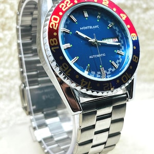 Könnte beinhalten: Eine automatische Montblanc Armbanduhr mit blauem Zifferblatt und rot-blauer Lünette. Die Uhr hat ein silberfarbenes Edelstahlarmband und -gehäuse. Das Zifferblatt zeigt das Datum und das Wort "AUTOMATIC".