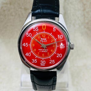 Może przedstawiać: Zegarek naręczny w stylu vintage z czerwoną tarczą, srebrną kopertą i czarnym skórzanym paskiem. Tarcza zegarka posiada białe cyfry oraz napis "hmt PILOT" i "PARA SHOCK 17 JEWELS". Zegarek jest prezentowany na przezroczystym stojaku.