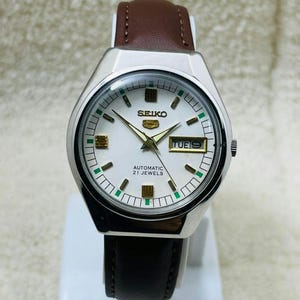 Op de afbeelding: Een zilverkleurig Seiko automatisch polshorloge met een witte wijzerplaat, gouden wijzers en een bruine leren band. De wijzerplaat toont de dag en datum, met het woord "SEIKO" bovenaan. Het horloge heeft een zwarte leren band en een bruine leren band.