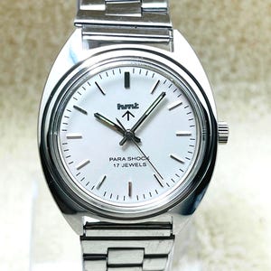 May include: A silver-toned wristwatch with a white face. The watch features black hands and markers, with the text "PARA SHOCK 17 JEWELS" and a logo. The watch has a silver band and a crown on the side.