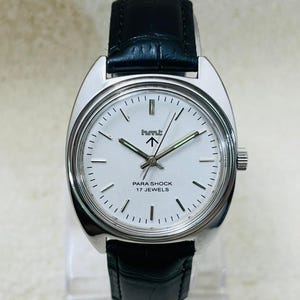 May include: A silver-toned wristwatch with a white face and black leather band. The watch face features black hour markers, a logo, and the text "PARA SHOCK 17 JEWELS". The watch has a silver bezel and a black leather strap.