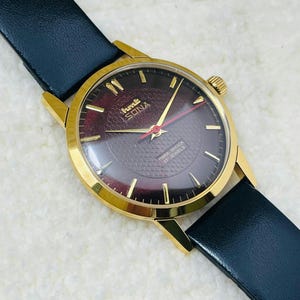 Könnte beinhalten: Eine goldfarbene Armbanduhr mit burgunderfarbenem Zifferblatt und schwarzem Lederarmband. Das Zifferblatt der Uhr hat goldene Zeiger und Markierungen mit dem Text "hmt SONA" und "PARA SHOCK 17 JEWELS". Die Uhr hat ein klassisches, Vintage-Design.