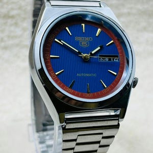 May include: A silver Seiko automatic wristwatch with a blue dial and a red inner ring. The watch has gold hands and markers, and a date display. The watchband is made of silver metal links.