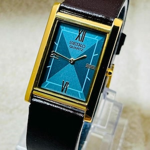 May include: A rectangular Seiko Quartz wristwatch featuring a gold-tone case and a dark teal dial with a geometric pattern. The dial displays Roman numerals at the twelve and six o'clock positions, gold-tone hands, and a black leather strap.