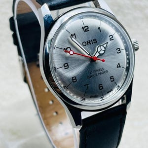 May include: Silver Oris watch with a black leather strap. The watch face features silver radial lines, black numerals, and a red second hand. The watch is marked "17 JEWELS SHOCK PROOF SWISS MADE".