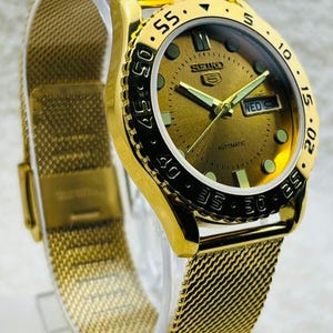 Pode incluir: Um relógio de pulso automático Seiko 5 dourado com uma pulseira de malha. O mostrador do relógio é dourado com algarismos e ponteiros pretos. A moldura tem marcações pretas. O relógio é exibido em um suporte transparente.