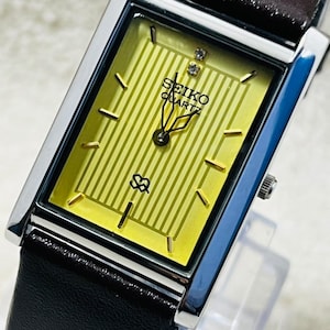 May include: A rectangular Seiko Quartz wristwatch featuring a yellow dial with vertical stripes, gold-tone hour markers, and two small clear stones at the twelve o'clock position. The watch has a silver-tone metal case and a black leather strap.
