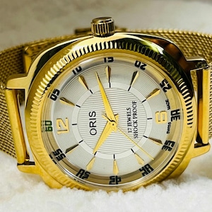 May include: A gold Oris wristwatch with a mesh strap. The watch face is silver with gold hands and markers. The text "ORIS" and "17 JEWELS SHOCK PROOF" are visible. A classic gold watch, a timeless accessory.