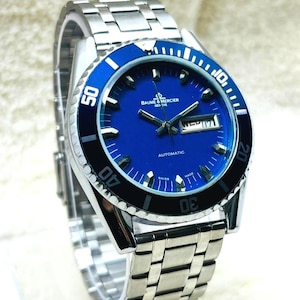 May include: A stainless steel Baume & Mercier Geneve automatic wristwatch. The watch has a blue dial with white markers, a date display, and a rotating bezel with black and white markings. The watch strap is also stainless steel.