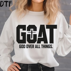 May include: White sweatshirt with the word "GOAT" in bold black letters, with a cross incorporated into the "O". Below the word "GOAT" is the phrase "GOD OVER ALL THINGS" in black. The sweatshirt has long sleeves.
