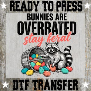May include: DTF transfer featuring a raccoon with a basket of Easter eggs. The text reads "READY TO PRESS BUNNIES ARE OVERRATED stay feral DTF TRANSFER". The raccoon is black and white, and the eggs are colorful.