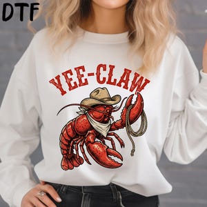 May include: White sweatshirt with a red graphic of a lobster wearing a cowboy hat and bandana, holding a rope. The text "YEE-CLAW" is above the lobster in red letters. The sweatshirt is a casual style.