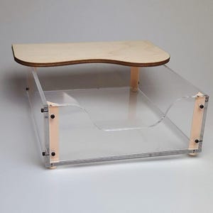 Peut inclure: Un organisateur de bureau en acrylique transparent doté d'une étagère supérieure en bois clair avec un bord avant incurvé. La structure est maintenue par des supports verticaux en bois aux coins, fixés par de petites vis métalliques noires.
