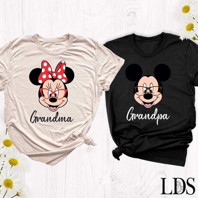 Puede incluir: Dos camisetas, una crema y otra negra, con personajes de dibujos animados. La camiseta crema tiene a Minnie Mouse con un lazo rojo y la palabra "Grandma". La camiseta negra tiene a Mickey Mouse con gafas y la palabra "Grandpa".