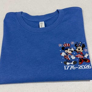 May include: A blue crew neck t-shirt featuring a graphic of two cartoon mice dressed in patriotic red, white, and blue clothing with star and stripe patterns. The design includes small white fireworks and the text 1776-2026 printed in white.