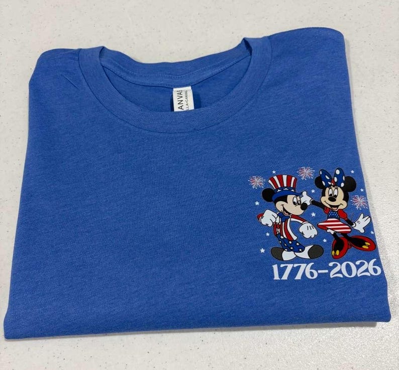 May include: A blue crew neck t-shirt featuring a graphic of two cartoon mice dressed in patriotic red, white, and blue clothing with star and stripe patterns. The design includes small white fireworks and the text 1776-2026 printed in white.