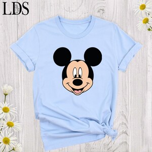 May include: Light blue t-shirt featuring a large graphic of Mickey Mouse's face. The cartoon character's face is black, white, and pink. The shirt has short sleeves and a crew neck. The letters "LDS" are in the upper left corner.