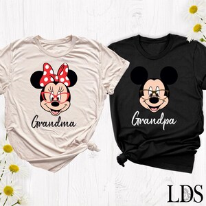 Puede incluir: Dos camisetas, una crema y otra negra, con personajes de dibujos animados. La camiseta crema tiene a Minnie Mouse con un lazo rojo y la palabra "Grandma". La camiseta negra tiene a Mickey Mouse con gafas y la palabra "Grandpa".