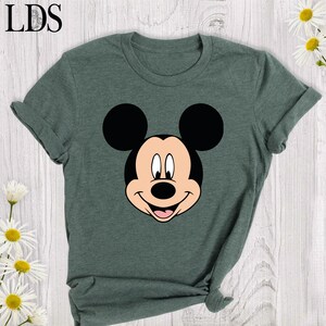 May include: A dark green t-shirt featuring a smiling Mickey Mouse face in black, white, and tan. The shirt has rolled-up sleeves and the letters "LDS" in the upper left corner. Daisies are in the background.