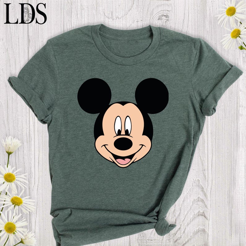 May include: A dark green t-shirt featuring a smiling Mickey Mouse face in black, white, and tan. The shirt has rolled-up sleeves and the letters "LDS" in the upper left corner. Daisies are in the background.