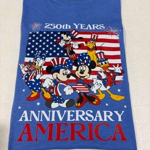 May include: A blue t-shirt featuring a graphic of Disney characters wearing patriotic red, white, and blue outfits in front of an American flag. The text reads 250th YEARS ANNIVERSARY AMERICA, accompanied by firework illustrations at the top corners.